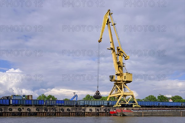 River port crane with clamshell or griper loading coal from open-top gondola car or wagon to river drag boats or barges moored by pier on cloudy day. Problems