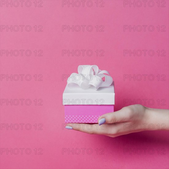 Female s hand holding box with white ribbon bow pink background