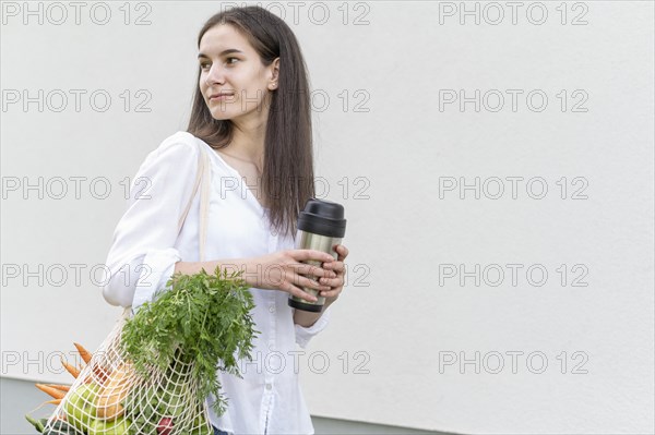 Mid shot woman holding reusable bag with groceries thermos outside