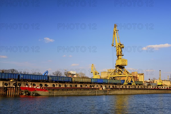 Colorful wide shot of river port crane loading open-top gondola cars on sunny day. Empty river drag boats or barges moored by pier. Empty cars ready for loading. Shipping raw materials