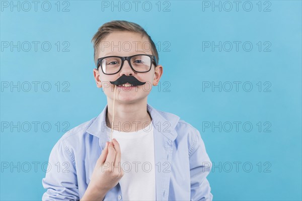 Boy wearing black eyeglasses holding black moustache prop front his upper lips