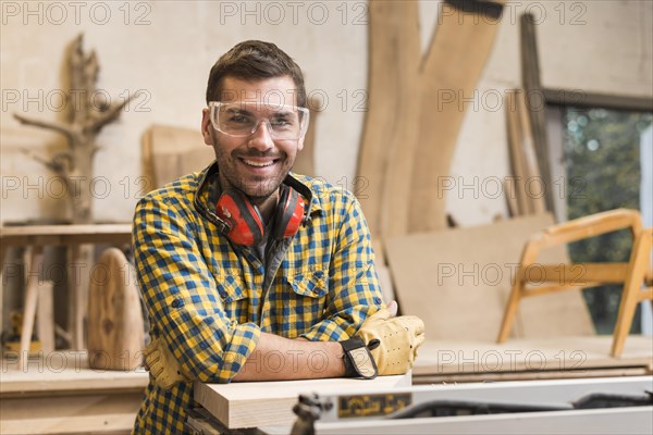 Smiling male carpenter with ear defender around her neck standing his workshop