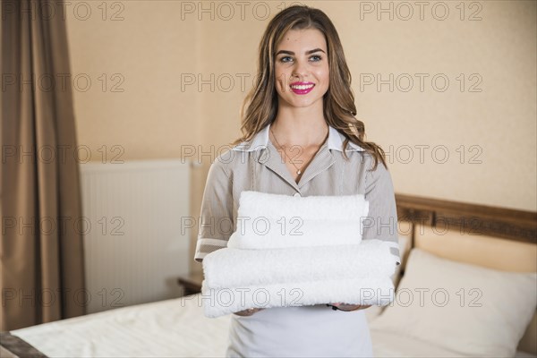 Smiling young maid holding white stack towels standing front bed