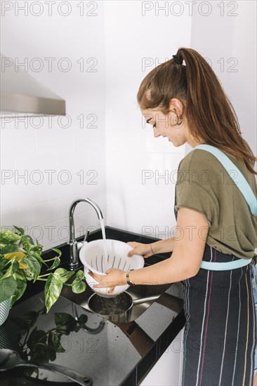 Smiling beautiful young woman holding colander flowing water sink