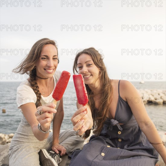 Smiling young women sitting seashore showing red popsicles