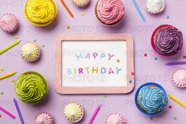 Wooden happy birthday white slate surrounded with muffins aalaw candles sprinkles pink background