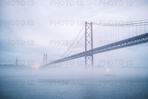 View of 25 de Abril Bridge famous tourist landmark of Lisbon connecting Lisboa and Almada in heavy fog mist wtih yacht boats passing under. Lisbon