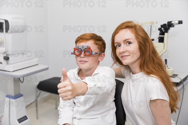 Boy with optometrist trial frame showing thumb up gesture sitting with young female ophthalmologist clinic