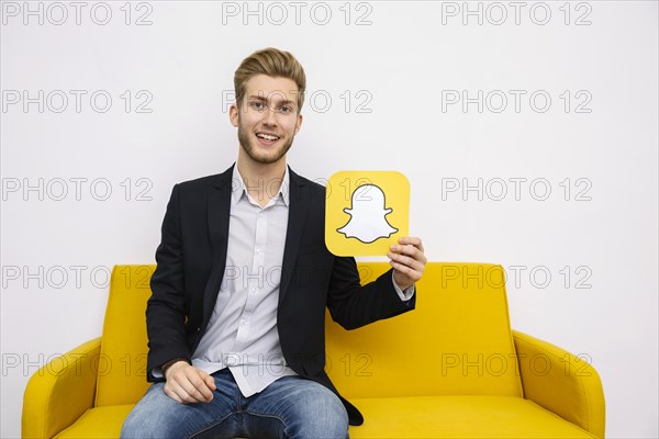 Portrait young man sitting yellow sofa holding snapchat icon