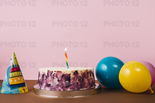 Colorful illuminated candle birthday cake with party hat balloons desk against pink background