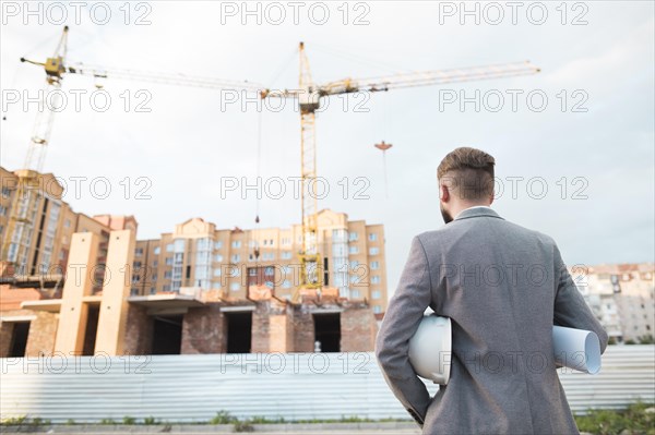 Rear view male architect holding blueprint hard hat looking construction site