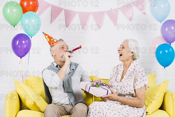 Happy woman with birthday gift looking her husband blowing party horn