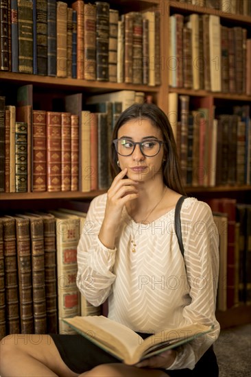 Pensive female student sitting with book library