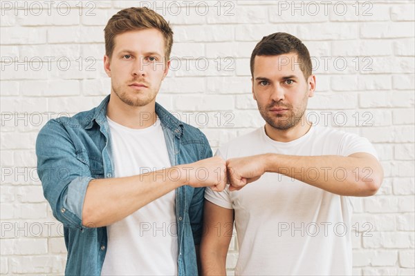Portrait two male friends bumping fist standing against white brick wall