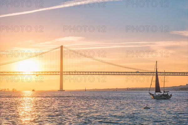 View of 25 de Abril Bridge famous tourist landmark of Lisbon connecting Lisboa and Almada over Tagus river with tourist yacht silhouette at sunset. Lisbon