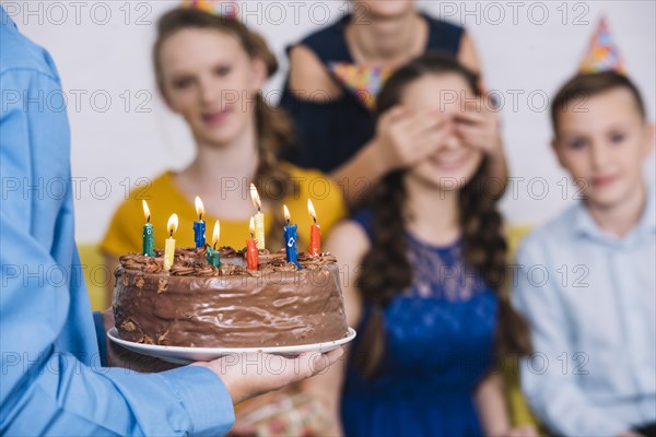 Close up boy s hand bringing chocolate cake birthday girl with covered eyes by her friend
