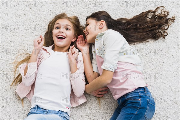 Excited girl listening friend whispering her ear lying carpet