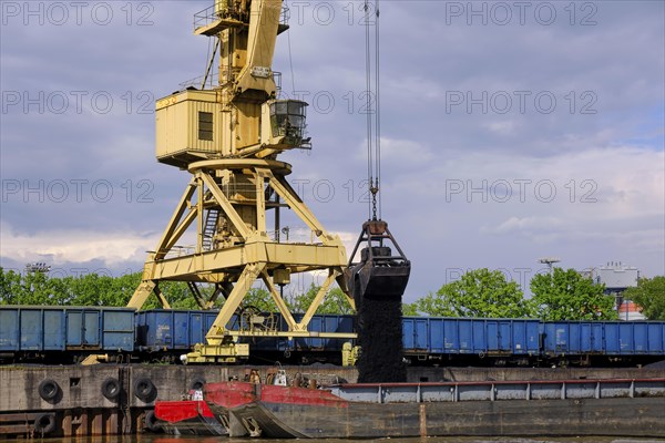 River port crane with clamshell or griper loading coal from open-top gondola car or wagon to river drag boats or barges moored by pier on cloudy day. Problems