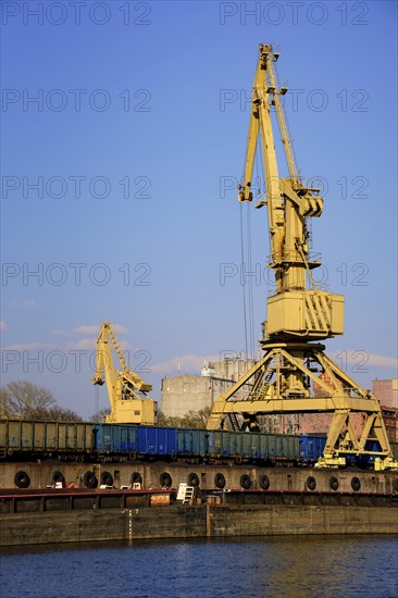 Colorful close vertical shot of river port crane loading open-top gondola cars on sunny day. Empty river drag boats or barges moored by pier. Empty cars ready for loading. Vertical image