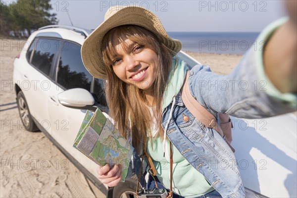 Smiling portrait female traveler holding map hand taking selfie with her car beach