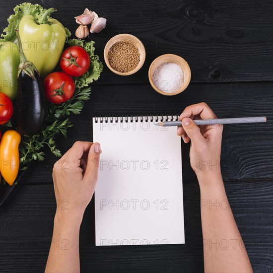 Human hand with spiral notepad pencil near vegetables spices black wooden backdrop