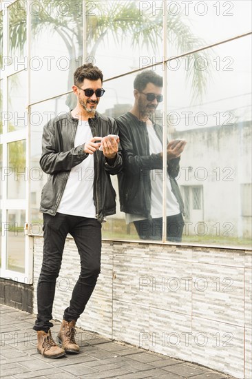 Handsome young man leaning glass wall using smartphone
