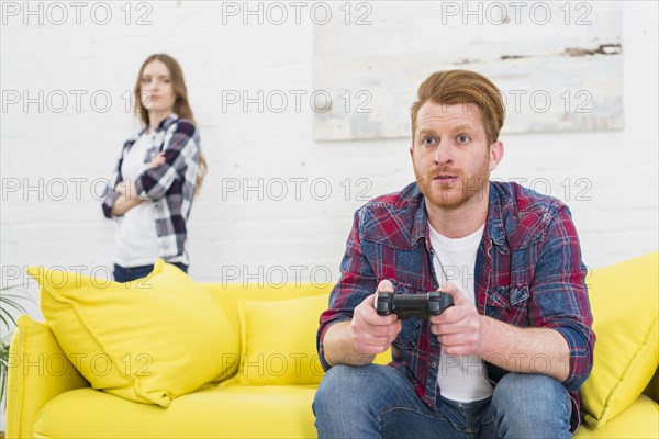 Serious young man playing game with video controller with her girlfriend standing background