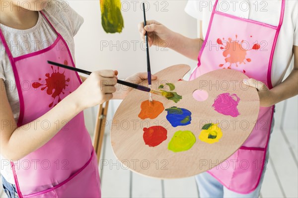 Elevated view two girls same pink apron mixing paint wooden palette with paint brush
