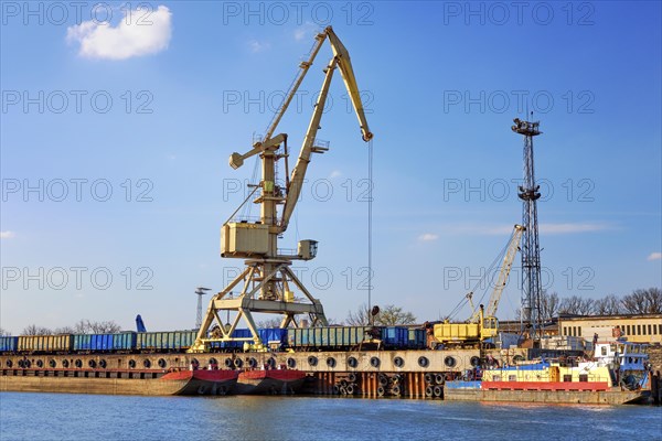 River port crane loading open-top gondola cars on sunny day. Empty river drag boats or barges moored by pier. Empty cars ready for loading. Colorful image