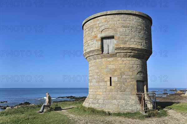 Old man sitting on bench looking at the sea and watchtower along the Cote Sauvage near Le Croisic