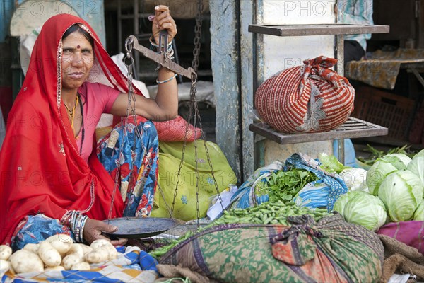 Sales women dressed in red sari weighing vegetables with balance at market in Bundi