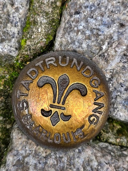 Brass-coloured metal medal embedded in a cobblestone street. The medal is circular and has a fleur-de-lis symbol in the centre. The medal is inscribed with the words Stadtrundgang and Saarlouis around the edge. Saarlouis