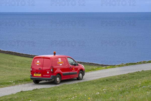 Postman driving red Peugeot Partner Royal Mail post van along desolate coastal road in Shetland