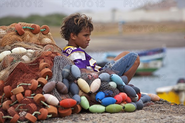 Young creole boy resting against pile of fishing nets in the harbour of fishing village Palmeira on the island of Sal