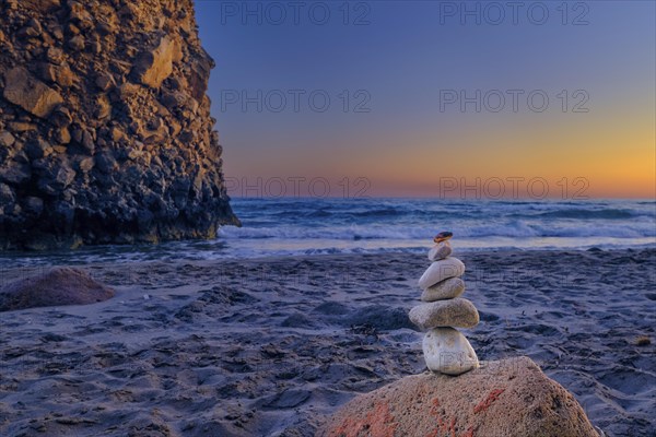 Small balance stone stack and big natural rock lit by sunset sun on sandy beach. Tidal waves in background
