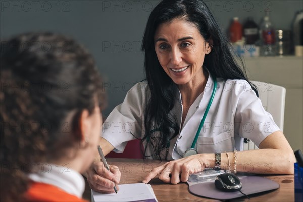 Smiling female doctor with stethoscope talking to woman in hospital in medical office and writing prescription