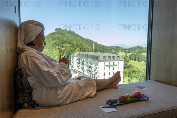 Woman in Bathrobe and Using Her SmartPhone and Enjoy the Window View over Mountain and Building in a Sunny Summer Day in Burgenstock