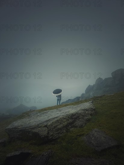 Rear view of a lonely man with umbrella stands on a rocky hill covered by haze. Moody and emotional scene with a lone stranger silhouette under the rain