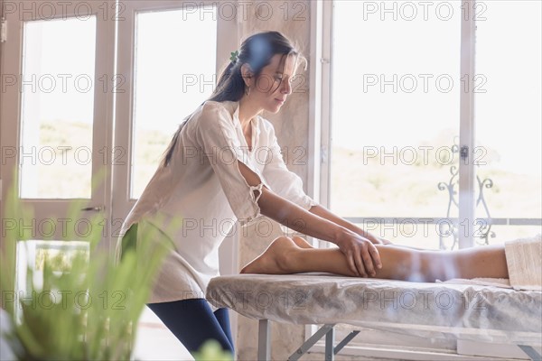 Portrait of a young masseuse performing a leg massage on a woman lying on a massage table. Side view of the professional masseuse. Luminous environment with incense