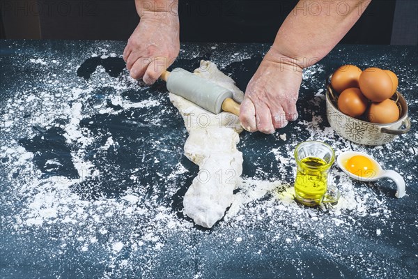 Woman kneading with a rolling pin a dough of flour to make bread on a black wooden table with olive oil
