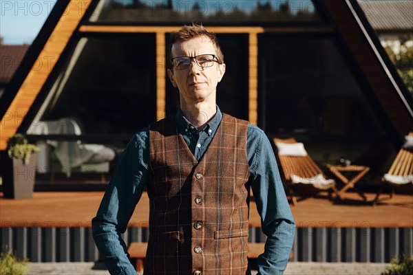 Outdoor portrait of middle aged man in jean shirt and wool vest with tiny house on background