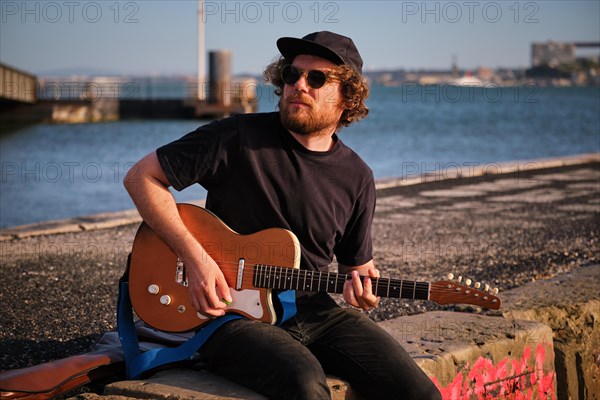 Hipster street musician in black playing electric guitar in the street sitting on pier embankment on sunset