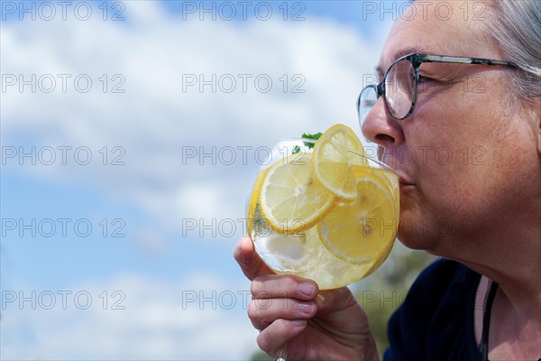 Woman seen in profile drinking a glass of gin and tonic with lemon slices and mint leaves with a blue sky with clouds in the background