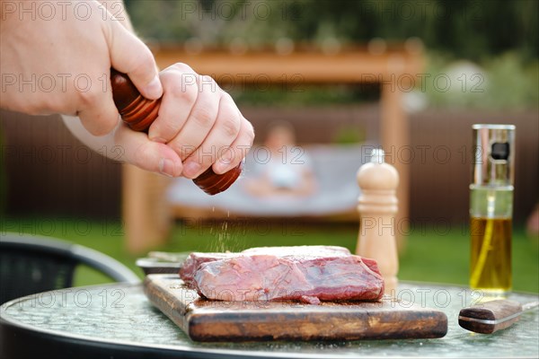 Side view of unrecognizable man adding pepper to raw beef steak making juicy meat on grill for dinner with his wife