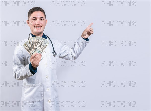 Happy latin doctor holding money and pointing to side. Handsome smiling doctor showing money and pointing side