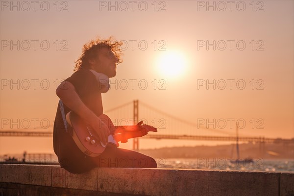 Hipster street musician in black playing electric guitar in the street on sunset on embankment with 25th of April bridge in background. Lisbon