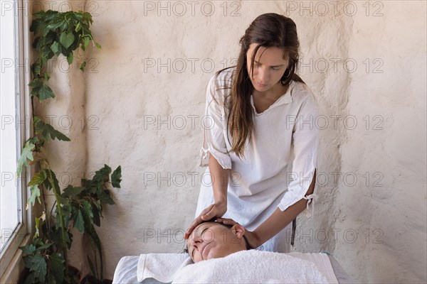 Beautiful young therapist performing massage in spa salon. Front view portrait of beautician doing facial massage to relaxed female client. Facial beauty treatment