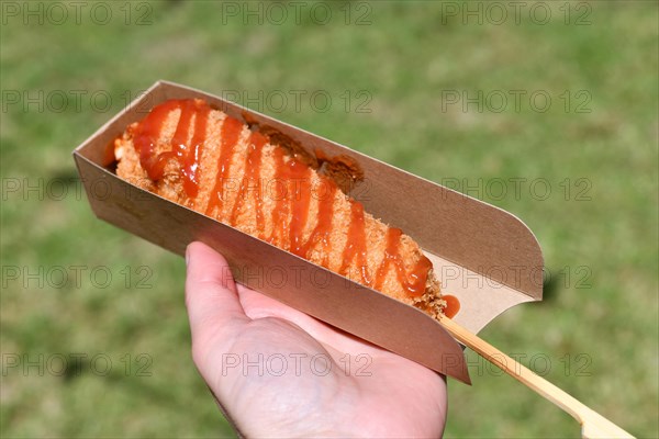 Hand holding Korean style corn dog coated in crispy panko breadcrumbs with hot sauce