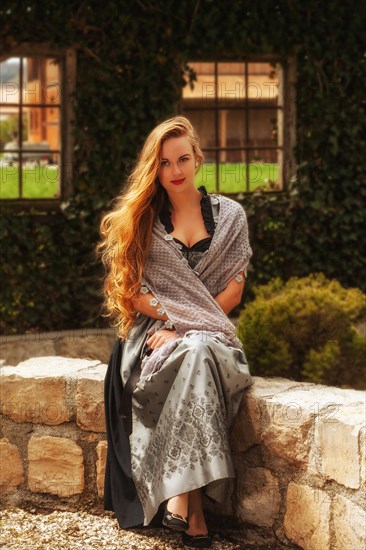 Woman with long hair and festive dirndl sits on a stone wall