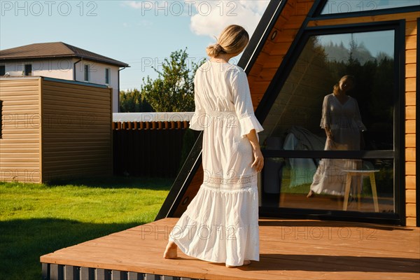 Playful woman in long white sundress looking at her reflection in the window
